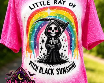 Pode incluir: Uma t-shirt rosa brilhante com efeito acid-wash apresentando um gráfico de uma morte de desenho animado segurando uma foice em frente a um arco-íris colorido. O texto diz LITTLE RAY OF PITCH BLACK SUNSHINE. Logotipo da Whiskey Witch Boutique.
