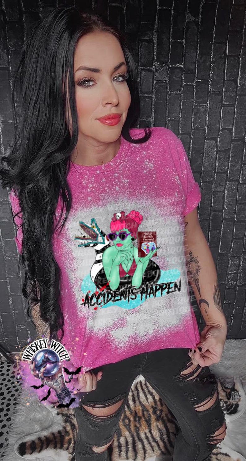 May include: A pink bleached t-shirt with a graphic print of a green-skinned woman with a black and white striped dress, sunglasses, and a book. The woman is sitting on a blue and white striped beach towel with a green alligator behind her. The text "Accidents Happen" is printed below the graphic.