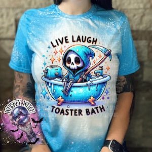 May include: A blue tie-dye t-shirt with a white bleach design featuring a cartoon skeleton in a blue hooded robe taking a bath in a blue bathtub with a toaster. The text "Live Laugh Toaster Bath" is printed on the shirt.