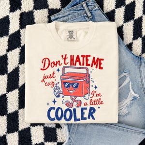 K&ouml;nnte beinhalten: Ein cremefarbenes Kurzarm-T-Shirt mit einer roten und blauen Grafik einer K&uuml;hlbox, die eine Sonnenbrille tr&auml;gt und ein Getr&auml;nk h&auml;lt. Der Text lautet: Don't HATE ME just cuz' I'm a little COOLER. Das Shirt hat ein Comfort Colors Markenetikett.