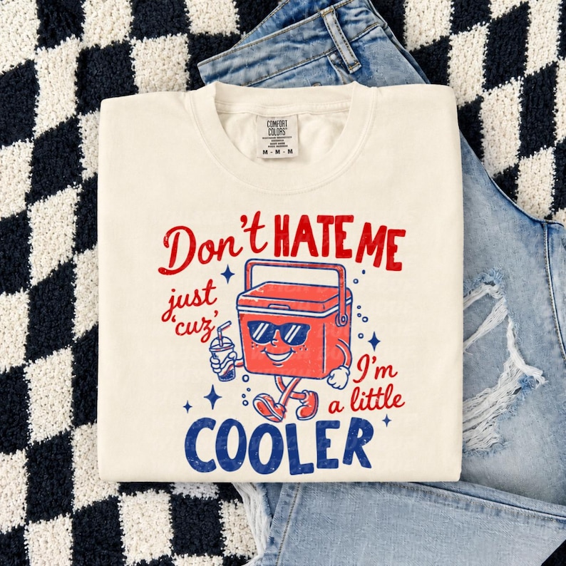K&ouml;nnte beinhalten: Ein cremefarbenes Kurzarm-T-Shirt mit einer roten und blauen Grafik einer K&uuml;hlbox, die eine Sonnenbrille tr&auml;gt und ein Getr&auml;nk h&auml;lt. Der Text lautet: Don't HATE ME just cuz' I'm a little COOLER. Das Shirt hat ein Comfort Colors Markenetikett.