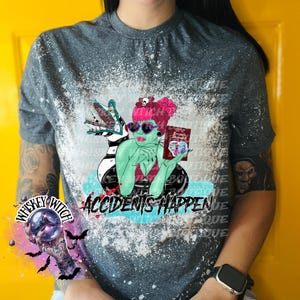 May include: A gray t-shirt with a bleach-splattered design featuring a green-skinned woman with a book that says "Recently Deceased" and a black and white striped creature. The text "Accidents Happen" is printed below the image. The shirt also has a small graphic of a crystal ball with the text "Whiskey Witch" around it.