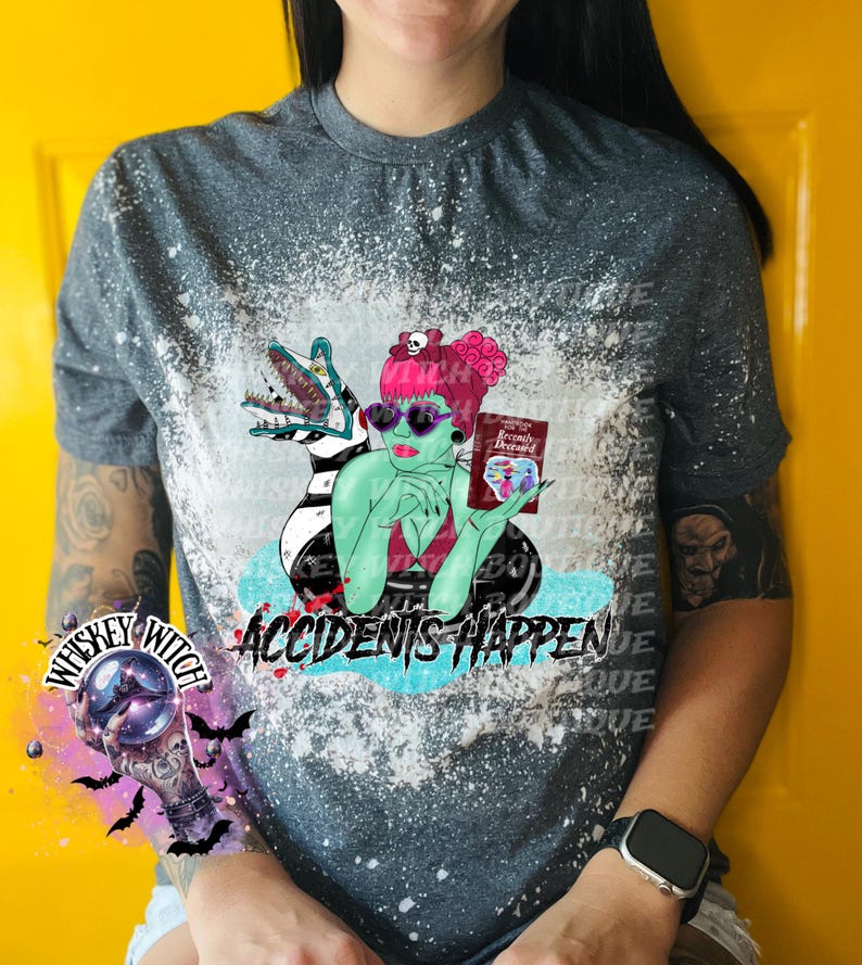May include: A gray t-shirt with a bleach-splattered design featuring a green-skinned woman with a book that says "Recently Deceased" and a black and white striped creature. The text "Accidents Happen" is printed below the image. The shirt also has a small graphic of a crystal ball with the text "Whiskey Witch" around it.