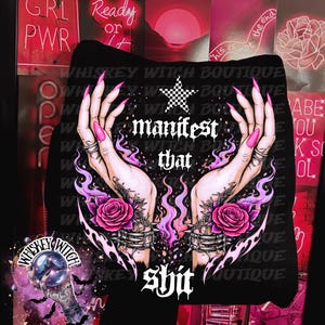 May include: Black hoodie featuring a graphic of hands with long pink nails, holding a star and roses, with the words "manifest that shit". The design includes flames and jewelry. The background has neon signs.