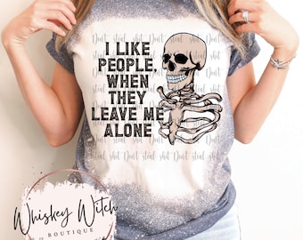 May include: A blue and white bleached t-shirt with the text "I LIKE PEOPLE, WHEN THEY LEAVE ME ALONE" and a skeleton graphic. The shirt also has the text "Whiskey Witch Boutique" in a circle.
