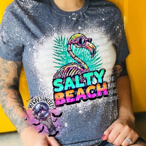May include: A blue t-shirt with a bleached design featuring a colorful flamingo wearing sunglasses. The flamingo graphic is above the words "SALTY BEACH" in vibrant colors. The shirt also has the text "Whiskey Witch Boutique".