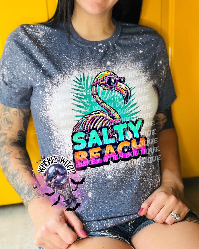 May include: A blue t-shirt with a bleached design featuring a colorful flamingo wearing sunglasses. The flamingo graphic is above the words "SALTY BEACH" in vibrant colors. The shirt also has the text "Whiskey Witch Boutique".