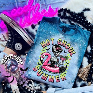 A Galapagos blue and bleach splattered effect  t-shirt with a graphic of a zombie woman in a flamingo pool float, holding a cocktail. The text "Hot Ghoul Summer" is printed on the shirt.