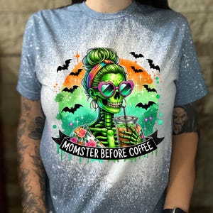 May include: Light blue t-shirt with a distressed design. The graphic features a green skeleton with heart-shaped sunglasses, drinking from a cup. The text "MOMSTER BEFORE COFFEE" is displayed on a banner, with bats and an orange and green background.