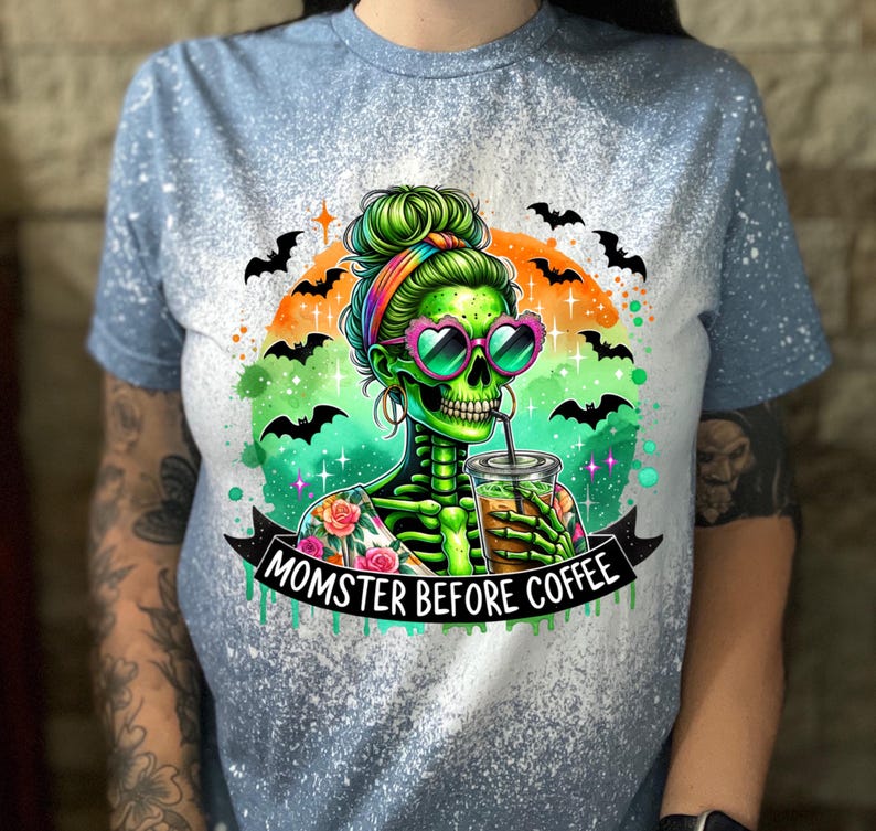 May include: Light blue t-shirt with a distressed design. The graphic features a green skeleton with heart-shaped sunglasses, drinking from a cup. The text "MOMSTER BEFORE COFFEE" is displayed on a banner, with bats and an orange and green background.