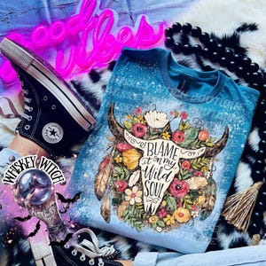 May include: A blue t-shirt with a bleached effect, featuring a floral bull skull design and the text "Blame it on my Wild Soul." The shirt is paired with black Converse sneakers and a black beaded necklace. A neon sign reads "Good Vibes."