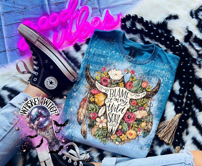 May include: A blue t-shirt with a bleached effect, featuring a floral bull skull design and the text "Blame it on my Wild Soul." The shirt is paired with black Converse sneakers and a black beaded necklace. A neon sign reads "Good Vibes."