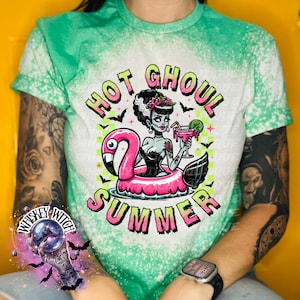 A green and bleach splattered effect  t-shirt with a graphic of a zombie woman in a flamingo pool float, holding a cocktail. The text "Hot Ghoul Summer" is printed on the shirt.