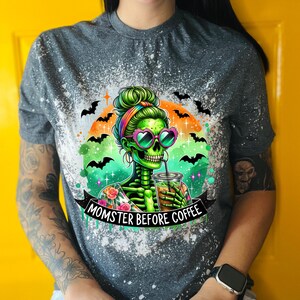 May include: Gray t-shirt with a graphic of a green skeleton woman wearing heart-shaped sunglasses, drinking coffee. The text "MOMSTER BEFORE COFFEE" is on a banner below the image. Bats and a rainbow arch the image.