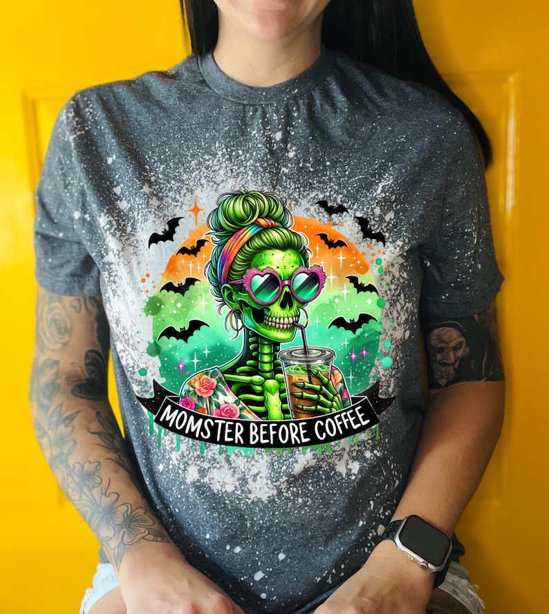 May include: Gray t-shirt with a graphic of a green skeleton woman wearing heart-shaped sunglasses, drinking coffee. The text "MOMSTER BEFORE COFFEE" is on a banner below the image. Bats and a rainbow arch the image.