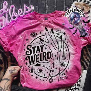 Handmade bleached styles shirt. Close-up of witchy Stay Weird shirt design with celestial artwork, mystical hands, stars, and bold black text