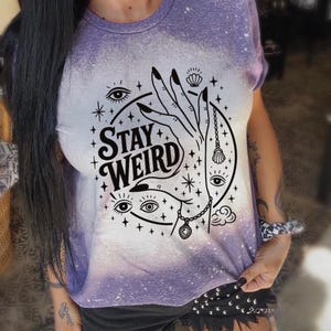 Handmade bleached styles shirt. Close-up of witchy Stay Weird shirt design with celestial artwork, mystical hands, stars, and bold black text