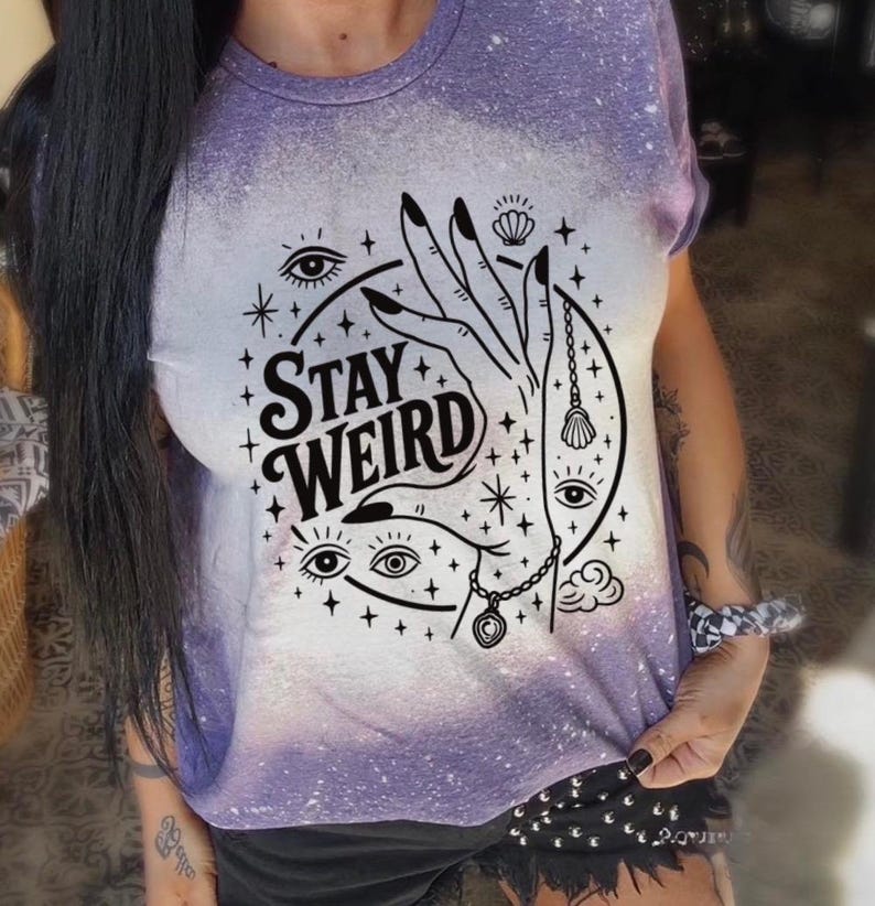 Handmade bleached styles shirt. Close-up of witchy Stay Weird shirt design with celestial artwork, mystical hands, stars, and bold black text