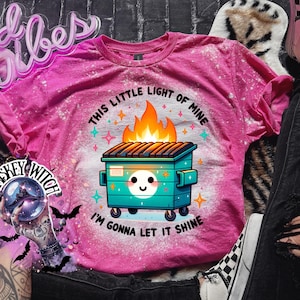 Pink t-shirt with a bleach-splattered design featuring a cartoon dumpster on fire with the text "This little light of mine I'm gonna let it shine".