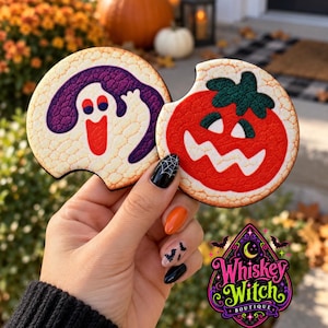 May include: Two round car coasters with a textured surface. One coaster features a white ghost with a purple outline and blue eyes. The other coaster features a red jack-o'-lantern with a green stem and white eyes and mouth, with a notch cut out.