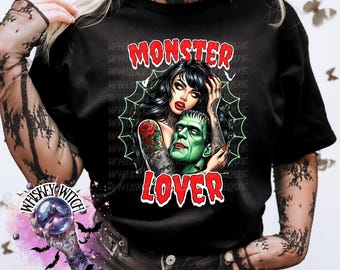 May include: Black t-shirt with a graphic featuring a woman with tattoos embracing Frankenstein's monster. The text "MONSTER LOVER" is in red, outlined in white. The design includes a spiderweb and the words "WHISKEY WITCH".