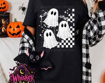 May include: A black t-shirt featuring a graphic print of three white ghosts with large black eyes, positioned above a black and white checkered pattern. The design includes small white star accents and the text Whiskey Witch Boutique.