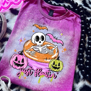 May include: A purple t-shirt with a Halloween-themed graphic. The design features a skeleton in a pool float, bats, and jack-o'-lanterns. The text "Just floatin'" is written in a playful font. The shirt has a bleached, distressed look.
