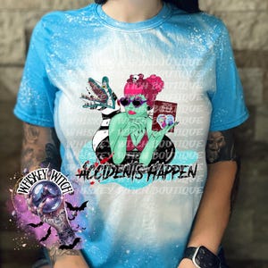 May include: A blue tie-dye t-shirt with a white bleach design featuring a green monster with a pink-haired woman sitting on its back. The woman is holding a book that says "Mommy Dearest" and the text "Accidents Happen" is below the image.