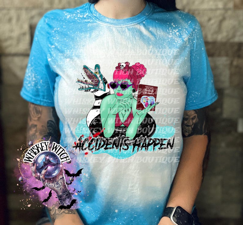 May include: A blue tie-dye t-shirt with a white bleach design featuring a green monster with a pink-haired woman sitting on its back. The woman is holding a book that says "Mommy Dearest" and the text "Accidents Happen" is below the image.