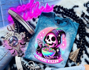 May include: A teal blue t-shirt with a tie-dye effect and a graphic of a skeleton wearing a hooded robe and eating cereal. The text "Cereal Killer" is printed below the graphic.