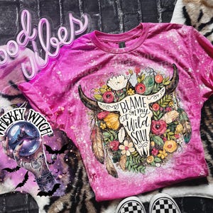 May include: A pink t-shirt with a bleached design featuring a bull skull adorned with flowers and the text "Blame it on my Wild Soul." The shirt has short sleeves and a crew neck.