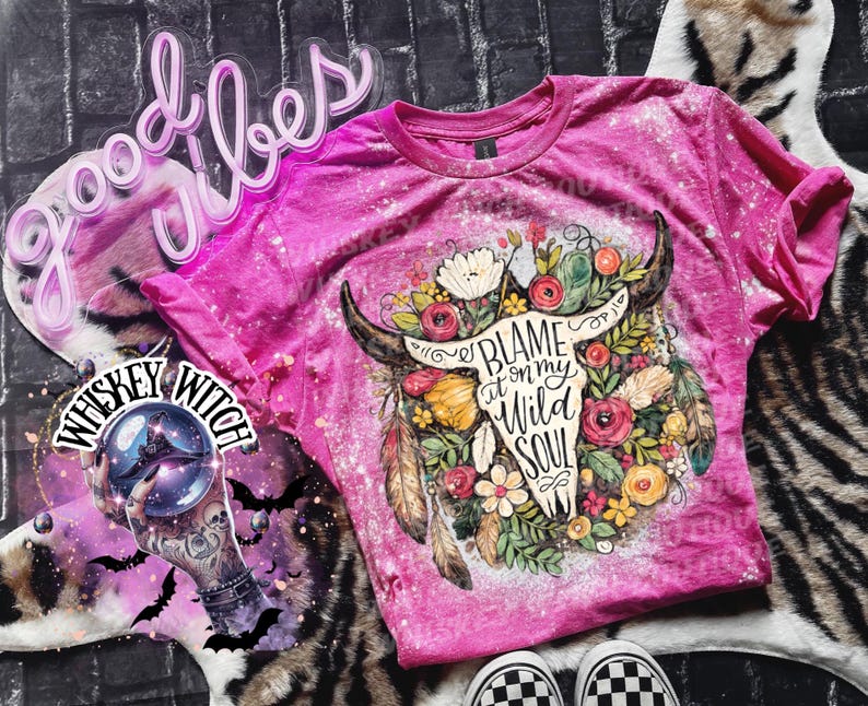 May include: A pink t-shirt with a bleached design featuring a bull skull adorned with flowers and the text "Blame it on my Wild Soul." The shirt has short sleeves and a crew neck.