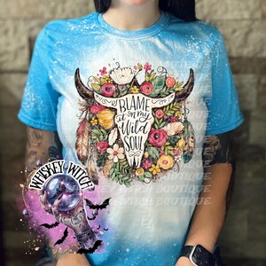 May include: A blue tie-dye t-shirt featuring a floral bull skull design with the text "BLAME at on my WILD SOUL". The design includes flowers, feathers, and horns. The shirt also has a "Whiskey Witch" logo.