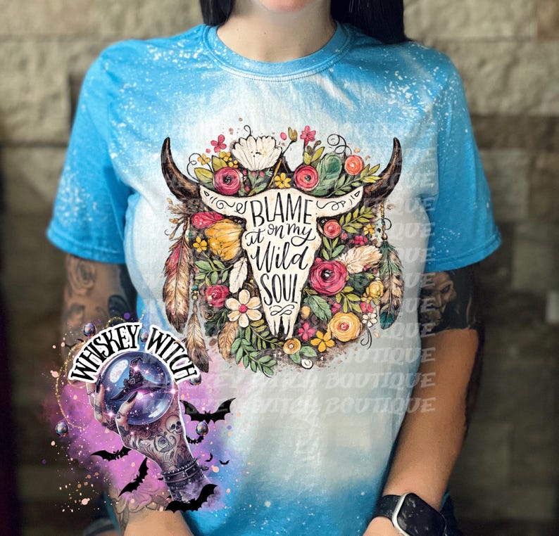 May include: A blue tie-dye t-shirt featuring a floral bull skull design with the text "BLAME at on my WILD SOUL". The design includes flowers, feathers, and horns. The shirt also has a "Whiskey Witch" logo.