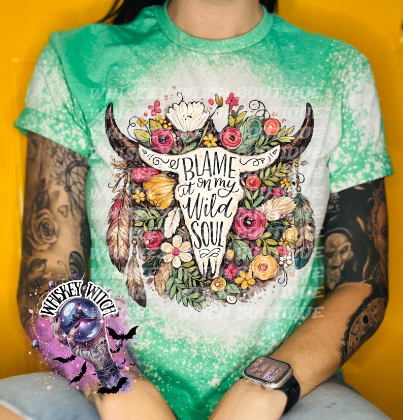 May include: A green and white t-shirt with a bleached design. The shirt features a floral skull graphic with the text "BLAME it on my WILD SOUL". The design includes flowers, feathers, and a bull skull.