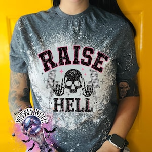 May include: Gray t-shirt with a distressed design featuring the text "RAISE HELL" in pink and black, a skull, and lightning bolts. The shirt has a crew neck and short sleeves.