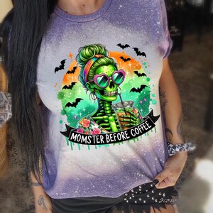 May include: Purple and white tie-dye t-shirt featuring a green skeleton with a bun, heart-shaped sunglasses, and a drink. The graphic includes bats and the text "MOMSTER BEFORE COFFEE". A fun Halloween-themed design.