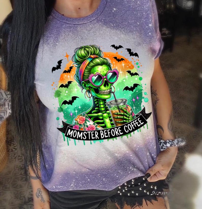 May include: Purple and white tie-dye t-shirt featuring a green skeleton with a bun, heart-shaped sunglasses, and a drink. The graphic includes bats and the text "MOMSTER BEFORE COFFEE". A fun Halloween-themed design.