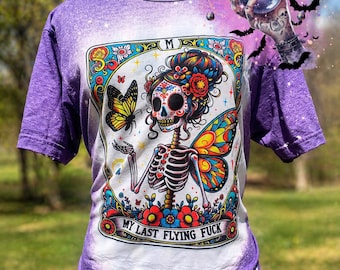 May include: A purple and white t-shirt featuring a colorful Day of the Dead-style skeleton woman with butterfly wings and a butterfly. The shirt has the text "MY LAST FLYING FUCK" in a banner at the bottom.