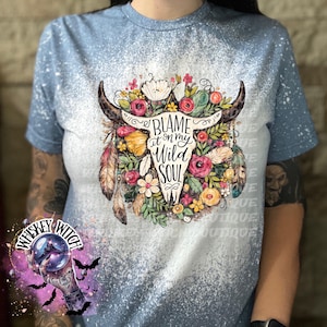 May include: Light blue t-shirt with a bleached design. The shirt features a graphic of a bull skull adorned with flowers, feathers, and the text "Blame it on my Wild Soul". The shirt is a casual wear item.