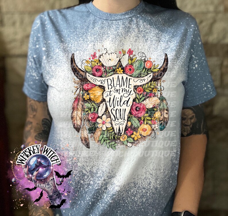 May include: Light blue t-shirt with a bleached design. The shirt features a graphic of a bull skull adorned with flowers, feathers, and the text "Blame it on my Wild Soul". The shirt is a casual wear item.