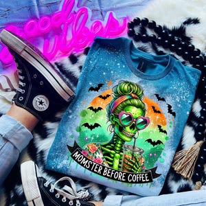 May include: A blue t-shirt with a graphic of a green skeleton wearing heart-shaped sunglasses and holding a drink. The shirt has the text "Momster Before Coffee" and is decorated with bats. Black Converse sneakers are also visible.