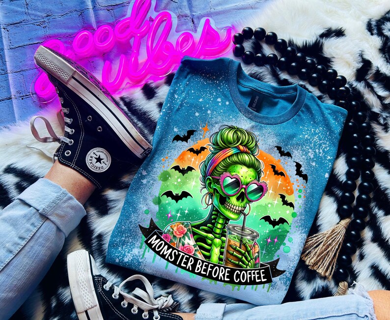 May include: A blue t-shirt with a graphic of a green skeleton wearing heart-shaped sunglasses and holding a drink. The shirt has the text "Momster Before Coffee" and is decorated with bats. Black Converse sneakers are also visible.