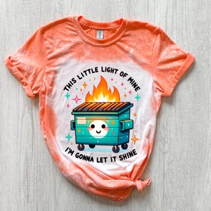 Orange t-shirt with a bleach-splattered design featuring a cartoon dumpster on fire with the text "This little light of mine I'm gonna let it shine".