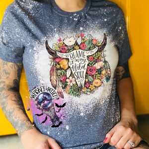 May include: A blue t-shirt with a bleached design featuring a bull skull surrounded by flowers and the text "Blame it on my Wild Soul." A smaller graphic of a crystal ball and bats is also visible.