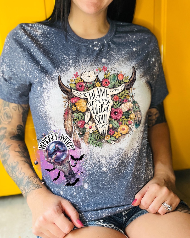 May include: A blue t-shirt with a bleached design featuring a bull skull surrounded by flowers and the text "Blame it on my Wild Soul." A smaller graphic of a crystal ball and bats is also visible.