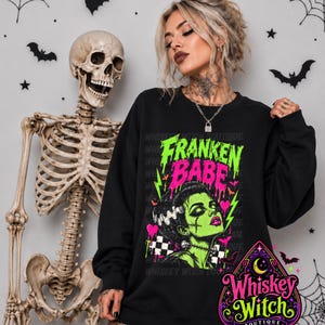 May include: A black long-sleeve sweatshirt featuring a graphic print with the text "FRANKEN BABE" in neon green and pink. The design includes a stylized illustration of a green-skinned character with dark hair, stitches, and bolts.