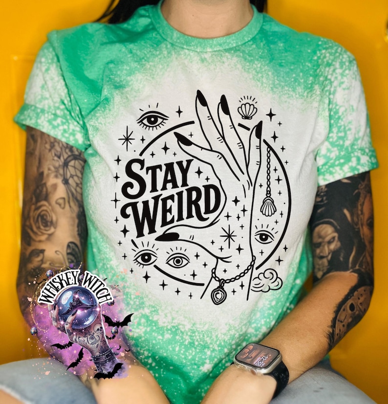 Handmade bleached styles shirt. Close-up of witchy Stay Weird shirt design with celestial artwork, mystical hands, stars, and bold black text