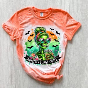 May include: Orange tie-dye t-shirt featuring a green skeleton with a colorful design, heart-shaped sunglasses, and a drink. The graphic includes bats and the text "MOMSTER BEFORE COFFEE".