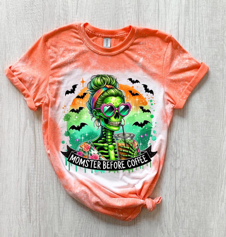 May include: Orange tie-dye t-shirt featuring a green skeleton with a colorful design, heart-shaped sunglasses, and a drink. The graphic includes bats and the text "MOMSTER BEFORE COFFEE".